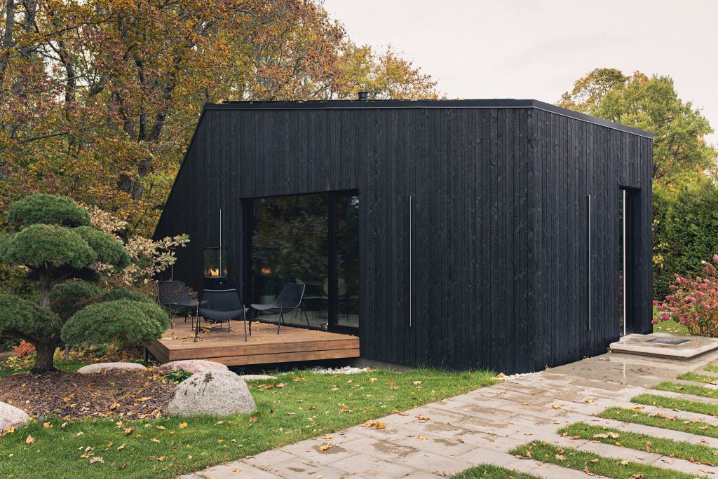 Casa moderna revestida con madera carbonizada de color negro intenso, rodeada de jardín con árboles y arbustos. La fachada minimalista destaca con grandes ventanales y una terraza de madera natural que crea contraste con el entorno verde y otoñal.