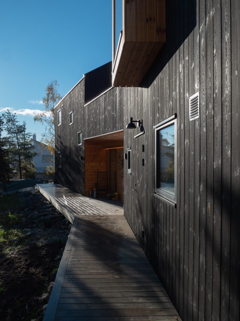 Fachada de vivienda moderna revestida con madera carbonizada en tonos oscuros, con pasarela de madera en la entrada y detalles de iluminación exterior. La combinación de texturas y la integración con el paisaje natural crean un diseño contemporáneo y acogedor.