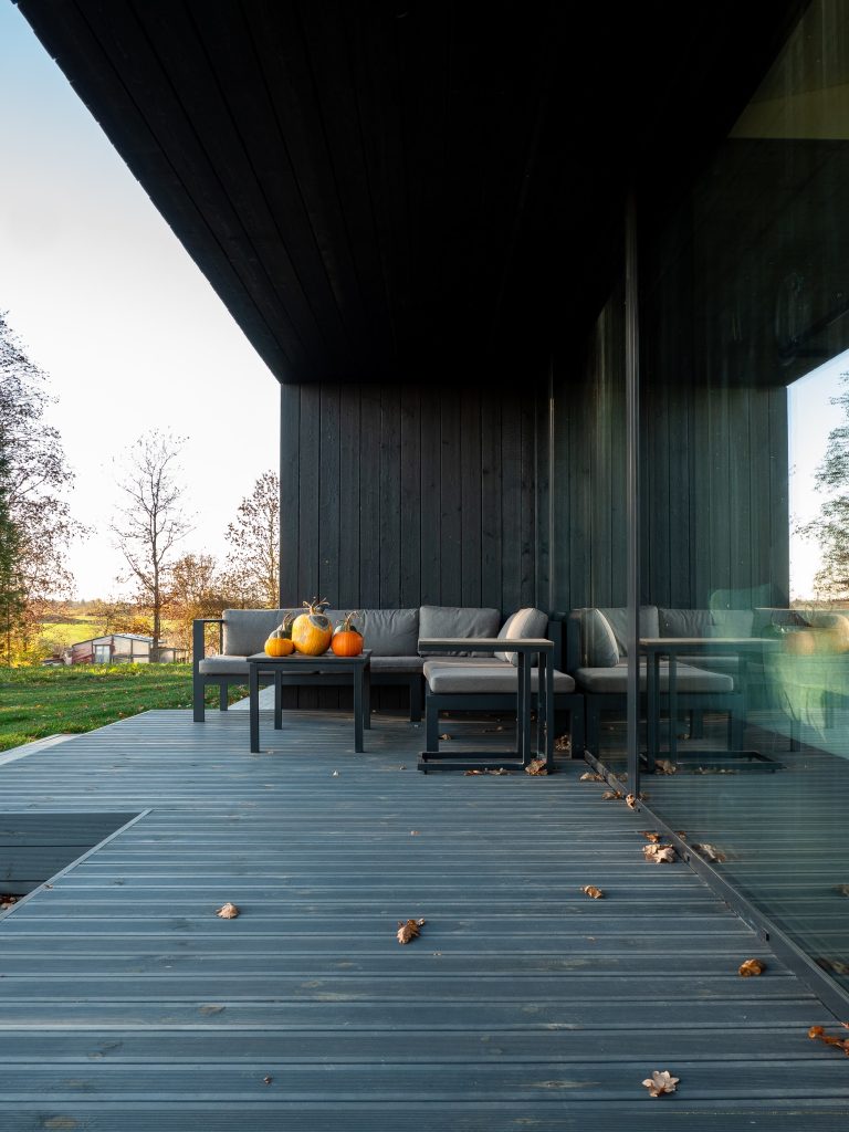 Terraza moderna con revestimiento de madera Yakisugi en acabado oscuro, sofá gris con cojines y mesa con calabazas decorativas frente a un ventanal de vidrio.