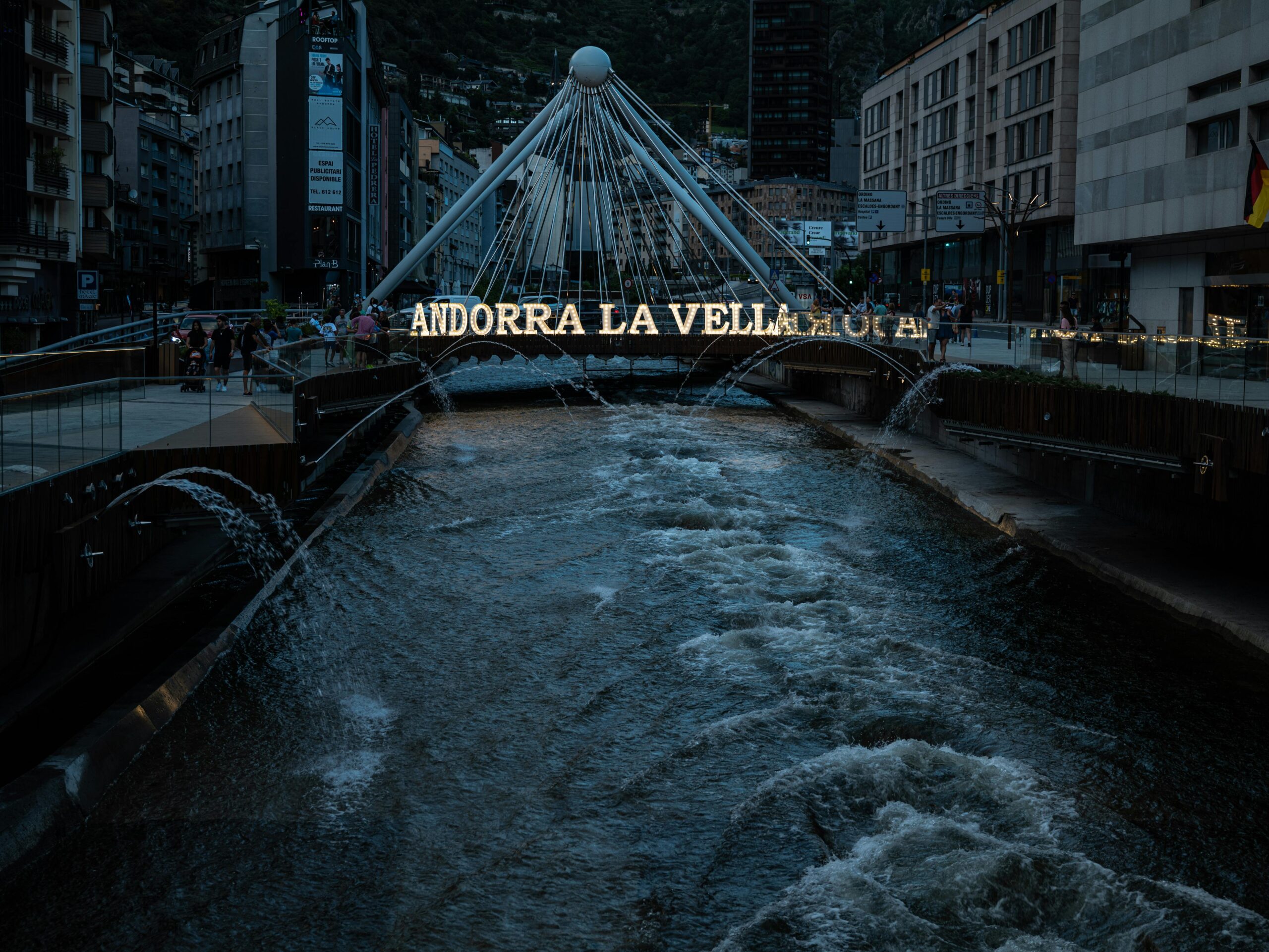 Vista del río en el centro de Andorra la Vella al atardecer, con el puente moderno y el letrero iluminado ‘Andorra la Vella’ sobre el agua.