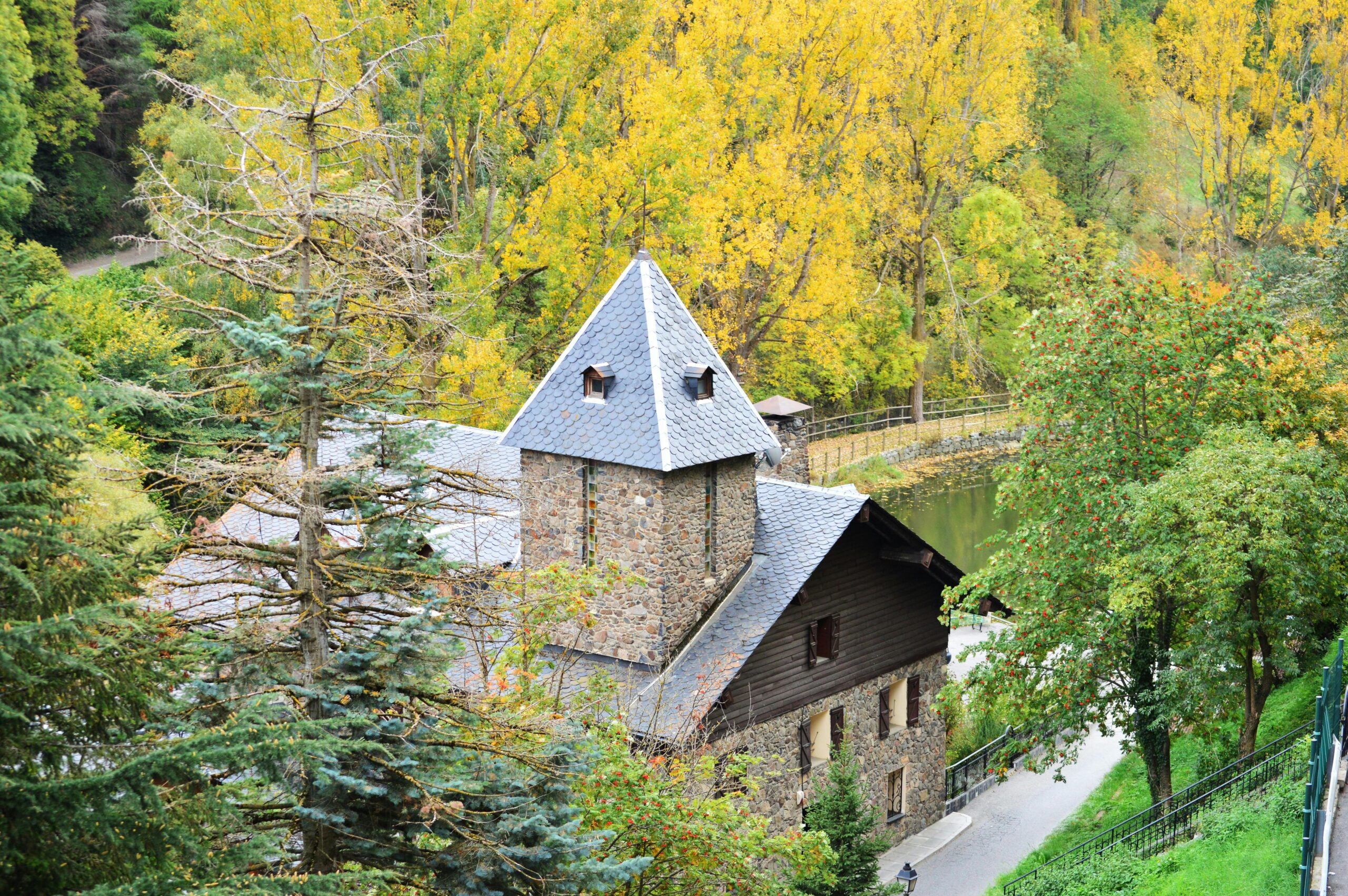 Casa tradicional de piedra con tejado de pizarra en Andorra, rodeada de árboles con hojas otoñales amarillas y verdes junto a un camino y un río.
