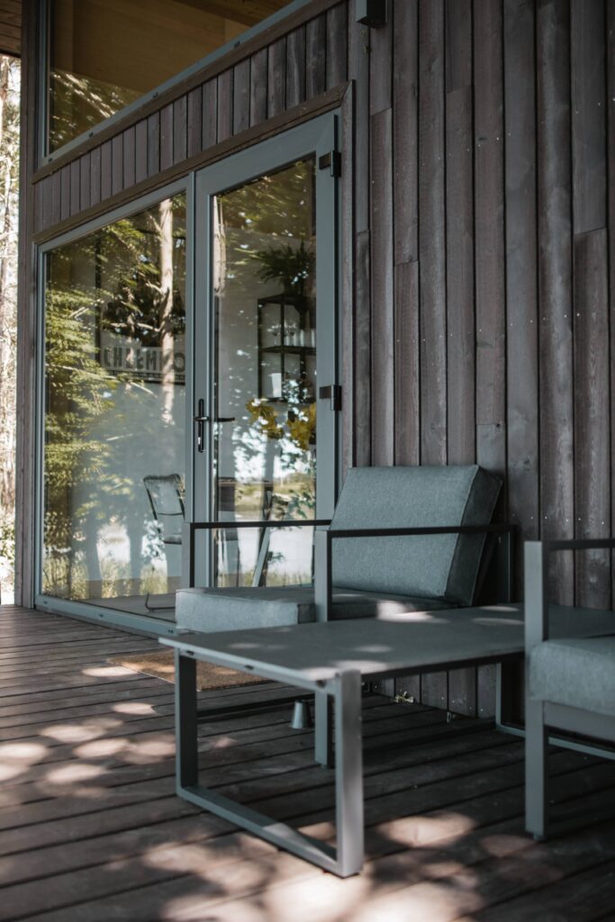 Detalle de terraza moderna con sillones grises y mesa metálica frente a una fachada de madera Yakisugi, mostrando la textura carbonizada y reflejos naturales en los ventanales de vidrio.