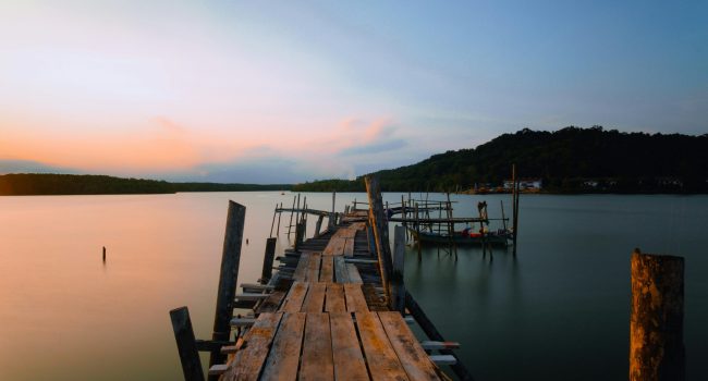 Cais de madeira rústico ao entardecer, sobre um rio tranquilo, com barcos ancorados e colinas cobertas de árvores ao fundo.