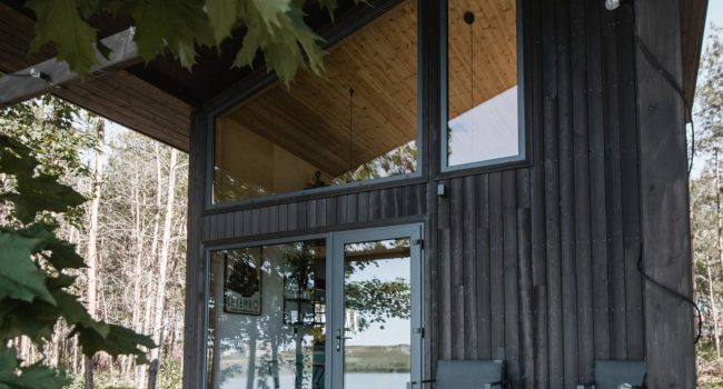 Fachada moderna de una casa de madera Yakisugi con acabado oscuro, grandes ventanales y terraza elevada frente al bosque, mostrando la textura carbonizada y el contraste natural con el entorno verde.