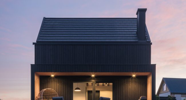 Casa contemporánea con fachada de madera carbonizada Yakisugi en tono oscuro, forma volumétrica simple, cubierta inclinada y un porche enmarcado iluminado al atardecer; paisaje de terreno rocoso en primer plano y cielo rosado al fondo.
