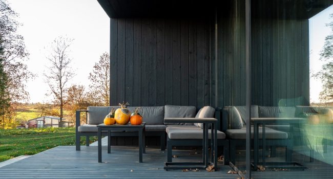 Terraza moderna con revestimiento de madera Yakisugi en acabado oscuro, sofá gris con cojines y mesa con calabazas decorativas frente a un ventanal de vidrio.